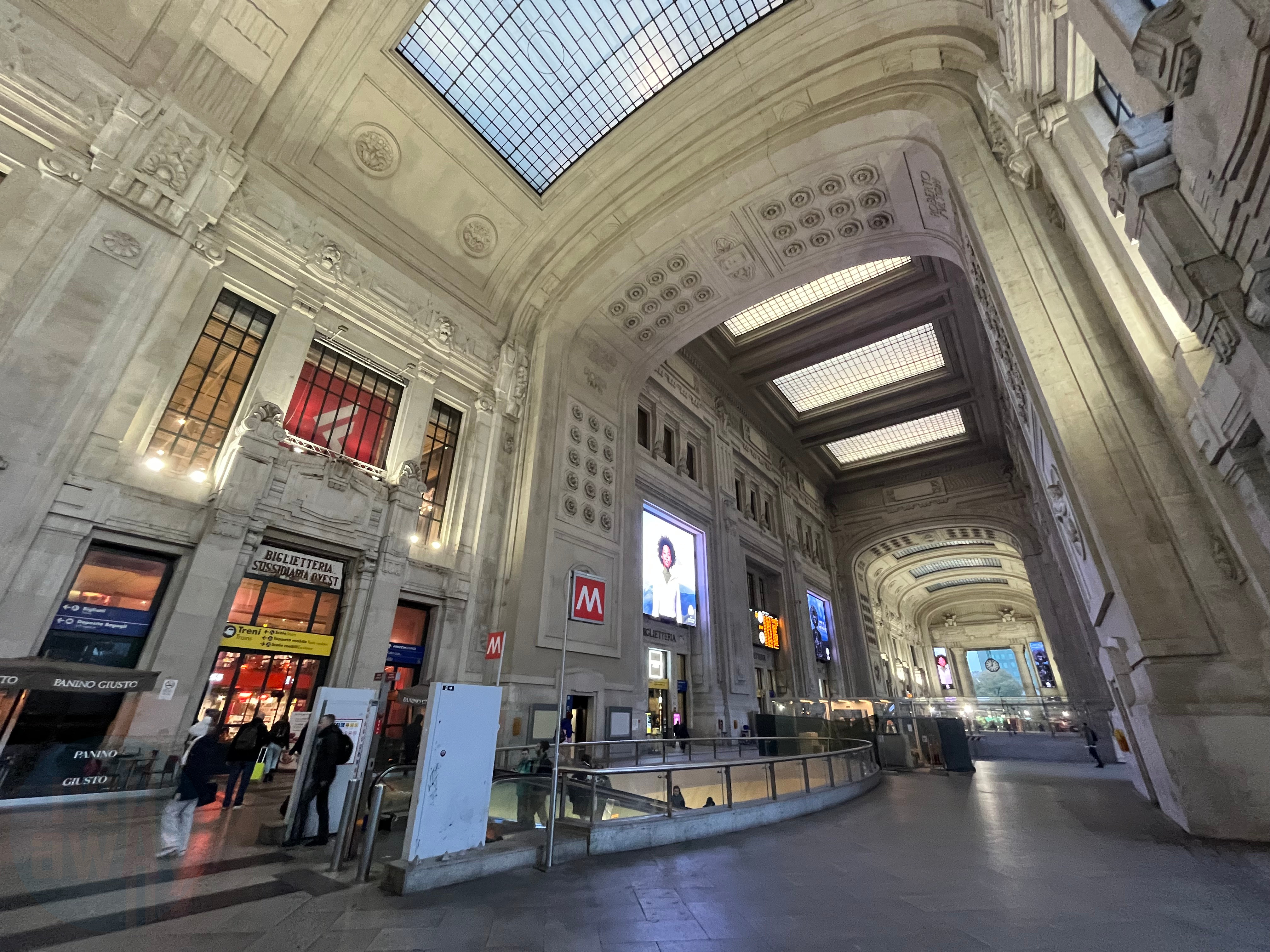 Main concourse of Milano Centrale station with its high ceiling. There is a ticket office on the left, escalators for the metro access