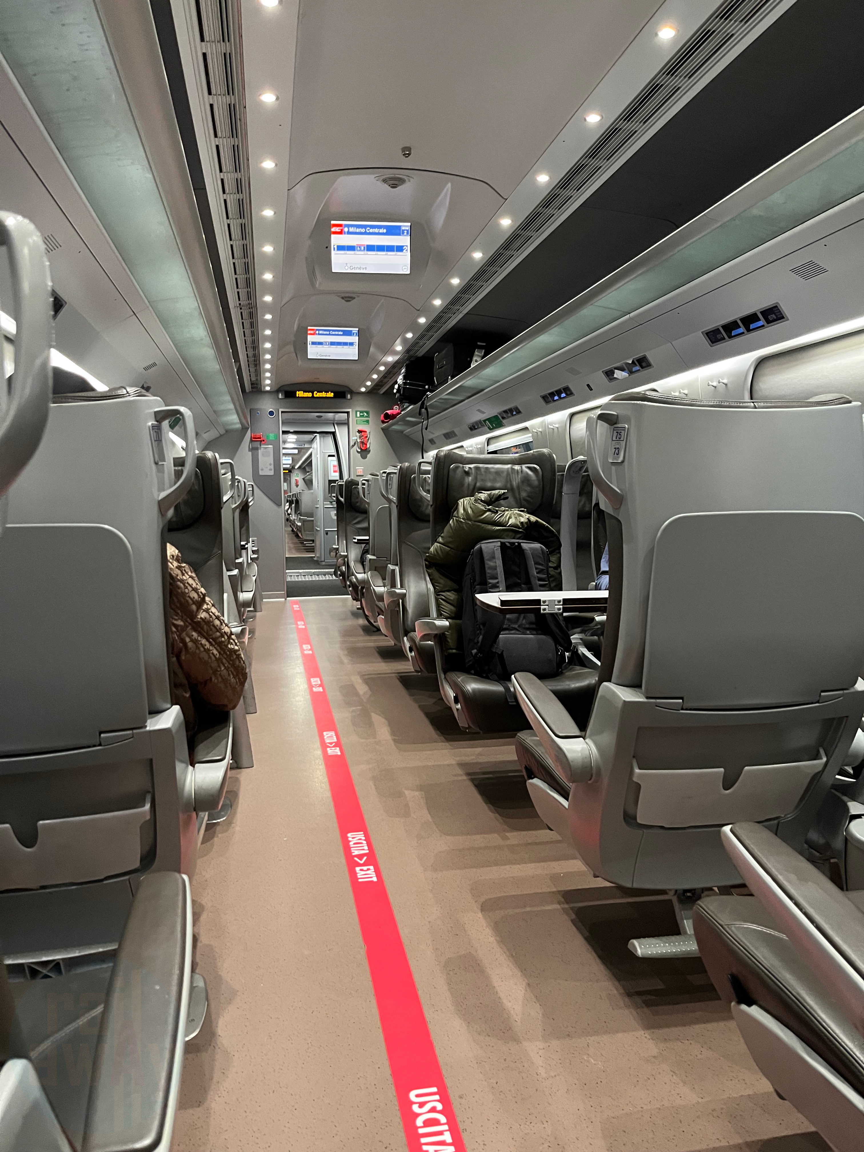 Brown leather seats in a 1+2 configuration in First Class with spot lights and vinyl flooring. There are digital screens displaying journey information.