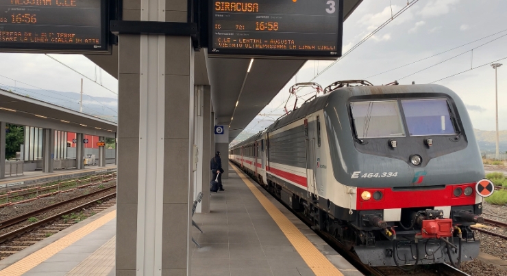 The mainline Trenitalia InterCity train at Giarre-Riposto station. Train is grey and white with a red stripe. There's a public information display board telling customers that the train goes to Siracusa and departs at 16:58.