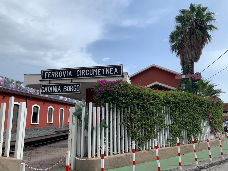 A photo of Catania Borgo station with red walls, a tree and a large sign saying Ferrovia Circumetnea