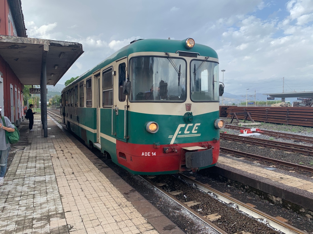 The green and white coloured FCE train at Giarre station that has a red terracotta station building